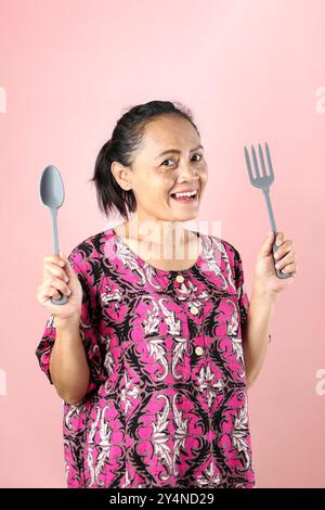 Femme d'âge moyen asiatique indonésienne tenant une cuillère et une fourchette, House Wives Core concept. Isolé sur fond rose Banque D'Images