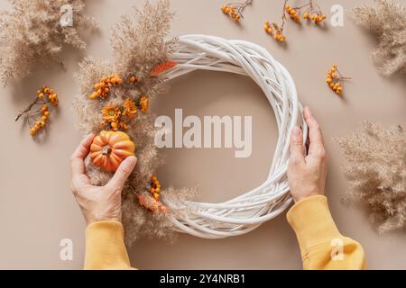 Femme en train de faire une couronne avec des fleurs orange et des matériaux naturels secs sur fond beige. Vue de dessus. Atelier pour la décoration artisanale de Thanksgiving. Banque D'Images