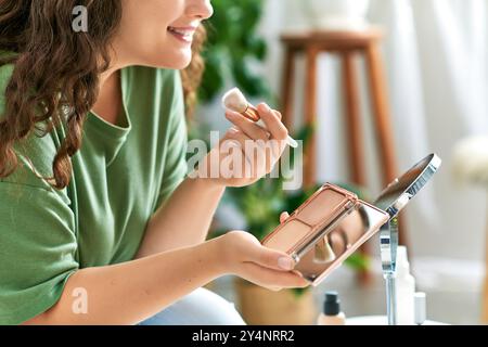Une jeune femme joyeuse applique le blush tout en étant entourée de ses produits de beauté préférés à la maison. Banque D'Images