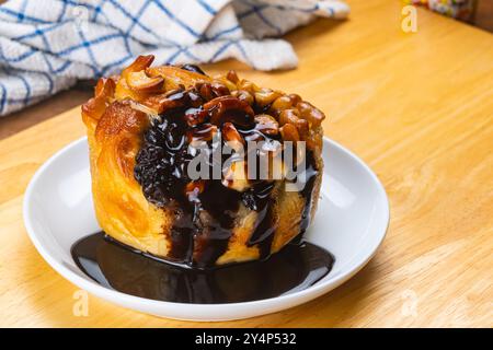 Vue en angle élevé de délicieux pain au beurre garni de noix et versant avec du sirop de chocolat dans un plat en céramique blanche sur planche de bois. Banque D'Images