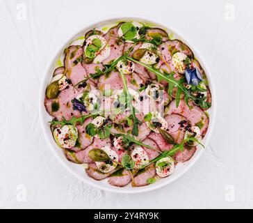 Tranches délicates de carpaccio de rôti de bœuf garni d'herbes, d'épices et de légumes verts sur une assiette blanche Banque D'Images