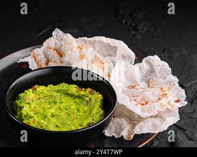 Guacamole vert vif dans un bol noir à côté de chips de riz croustillantes sur un fond texturé foncé Banque D'Images