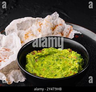 Guacamole vert vif dans un bol noir à côté de chips de riz croustillantes sur un fond texturé foncé Banque D'Images