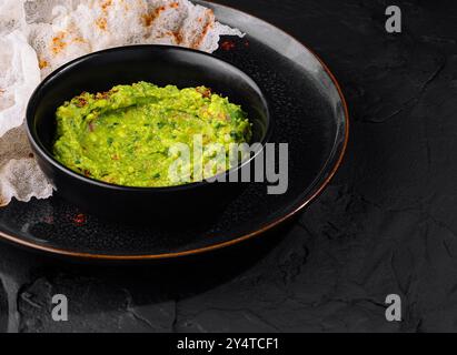 Guacamole vert vif dans un bol noir à côté de chips de riz croustillantes sur un fond texturé foncé Banque D'Images