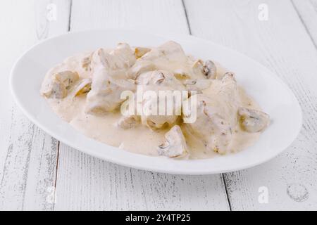 Poulet et champignons dans une sauce crémeuse, servis sur une assiette blanche sur une table en bois blanc Banque D'Images