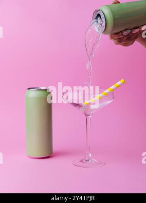 Femme verse une boisson rafraîchissante d'une canette dans un élégant verre avec une paille, sur un fond rose vif Banque D'Images