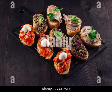 Délicieuse bruschetta avec tomates, mozzarella, pâté de viande et hareng couché sur une planche à découper en pierre noire Banque D'Images