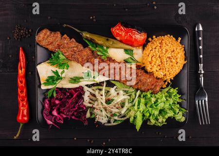 Traditionnel turc adana kebab servi sur une assiette avec tomates grillées, poivre, oignon, salade, persil, pain lavash et riz pilaf bulgur sur woo noir Banque D'Images