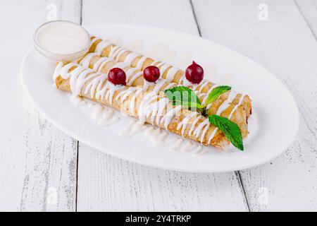 De délicieuses crêpes maison couchées sur une assiette blanche avec de la crème sure et des cerises attendent les clients sur une table en bois blanc Banque D'Images