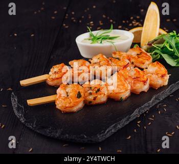 Brochettes de crevettes tigrées grillées servies sur une assiette en pierre avec herbes, citron et sauce blanche sur une table en bois noir Banque D'Images