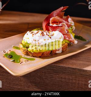 Toast avec de la purée d'avocat et oeuf poché décoré de prosciutto et de graines, arrosé d'huile d'olive Banque D'Images