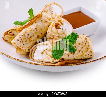 Crêpes fines avec fromage cottage et tranches de banane décorées de feuilles de menthe couchées sur une assiette blanche avec sauce sucrée Banque D'Images