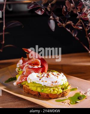 Toast avec de la purée d'avocat et oeuf poché décoré de prosciutto et de graines, arrosé d'huile d'olive Banque D'Images