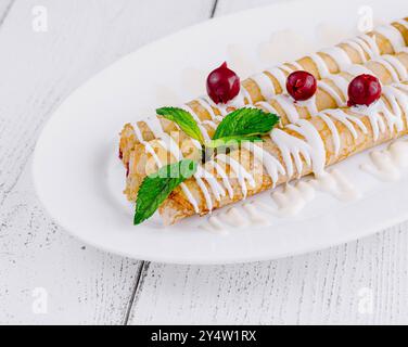 De délicieuses crêpes maison couchées sur une assiette blanche avec de la crème sure et des cerises attendent les clients sur une table en bois blanc Banque D'Images