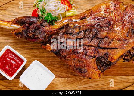 Le jarret d'agneau rôti est servi sur une planche de bois avec des légumes frais et des sauces Banque D'Images