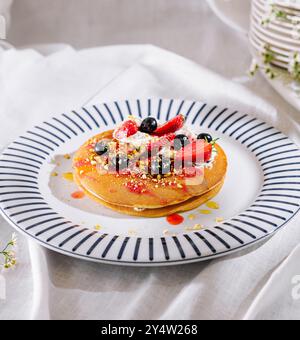 Délicieuse pile de crêpes couchées sur une assiette avec des rayures bleues, surmontées de fraises, de myrtilles, de pistaches et de crème fouettée, trempées de sirop Banque D'Images