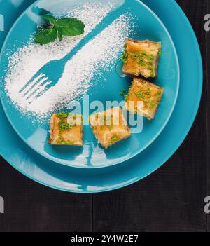 Délicieux baklava aux pistaches disposés sur une plaque bleue avec du sucre en poudre formant une fourchette, sur une table en bois noir Banque D'Images