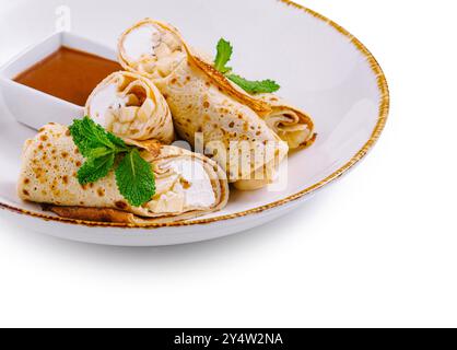 Crêpes fines avec fromage cottage et tranches de banane décorées de feuilles de menthe couchées sur une assiette blanche avec sauce sucrée Banque D'Images