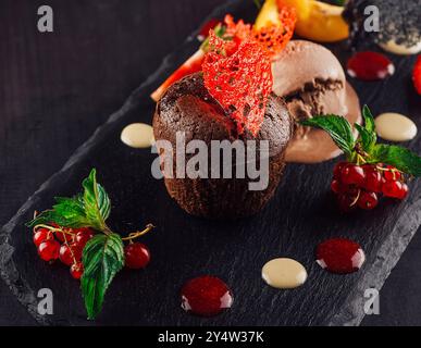 Gâteau de lave au chocolat servi sur une assiette en pierre noire avec une boule de glace, garnie de groseilles rouges, de menthe et d'une fraise Banque D'Images