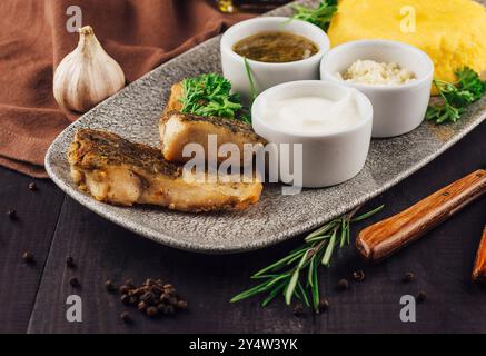 Filet de poisson frit servi sur assiette avec polenta, romarin, ail et sauces sur table en bois foncé Banque D'Images