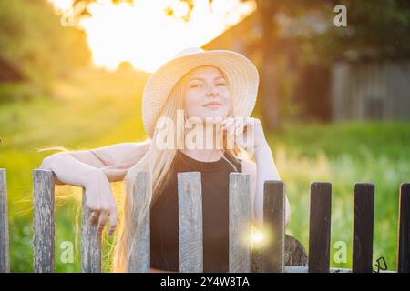 Femme blonde dans un chapeau de paille, se détendre par une clôture et admirer le coucher de soleil de la campagne Banque D'Images