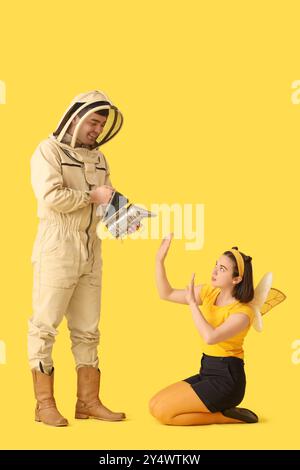 Apiculteur masculin avec fumeur et femme en costume d'abeille sur fond jaune Banque D'Images