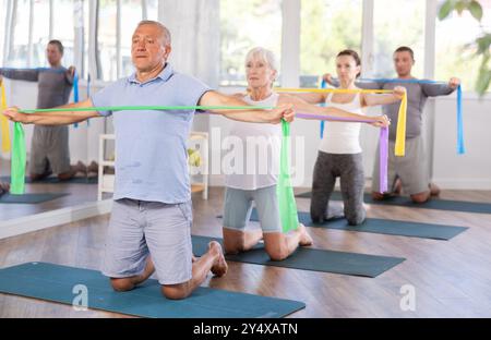 Groupe de personnes matures faisant des exercices d'étirement des bras en utilisant des bandes de résistance tout en s'agenouillant sur des tapis Banque D'Images