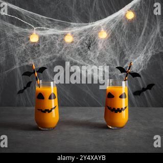 Boisson d'Halloween sur un fond gris avec des guirlandes lumineuses. Deux verres de jus avec un sourire effrayant et des pailles à boire avec des chauves-souris. Banque D'Images