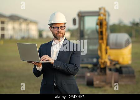 Directeur de construction en costume et casque sur un chantier de construction. Un employé ou un superviseur du directeur de la construction portant un casque de sécurité devant la maison Banque D'Images