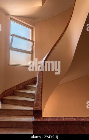 Un escalier en colimaçon torsadé avec des marches en marbre et bannister, avec des murs de couleur pêche, et une fenêtre fissurée montrant un ciel bleu. Banque D'Images