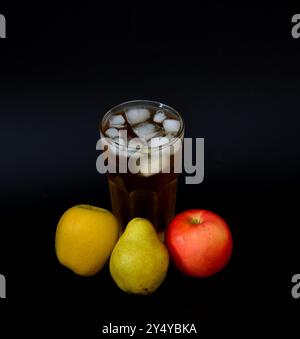 Un grand verre à facettes d'un mélange de jus de pomme et de poire de différentes variétés avec de la glace sur un fond noir, avec des fruits mûrs à proximité. Gros plan. Banque D'Images