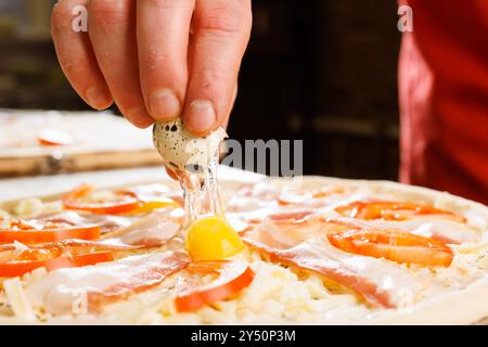 Gros plan d'une main cassant un œuf de caille sur une pizza garnie de jambon, de fromage et de tomates tranchées. La pizza est crue, mettant en valeur l'ingredien frais Banque D'Images