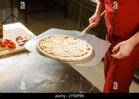 Un chef dans un uniforme rouge se prépare à placer une pizza avec du fromage et des tranches de tomate dans un four. Banque D'Images