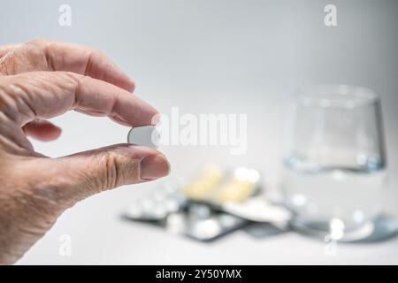 Main plus âgée tenant une pilule devant des paquets de médicaments flous et un verre d'eau, médecine et concept de soins de santé, fond gris clair, copie s. Banque D'Images