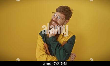 Un homme barbu rousse souriant dans des lunettes posant avec confiance sur un fond jaune, exsudant le charme décontracté. Banque D'Images