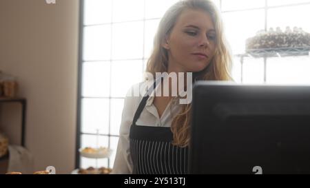 Jeune femme blonde dans une boulangerie portant un tablier, travaillant à l'intérieur derrière un comptoir avec des pâtisseries autour Banque D'Images