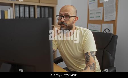 Bel homme hispanique avec une barbe et des tatouages travaillant à un bureau dans un cadre de bureau, profondément concentré sur un écran d'ordinateur dans un lieu de travail intérieur Banque D'Images