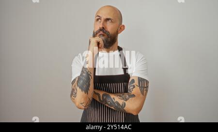 Homme hispanique avec une barbe et des tatouages portant un tablier sur un fond blanc semblant réfléchi tout en étant isolé sur un mur blanc. Banque D'Images
