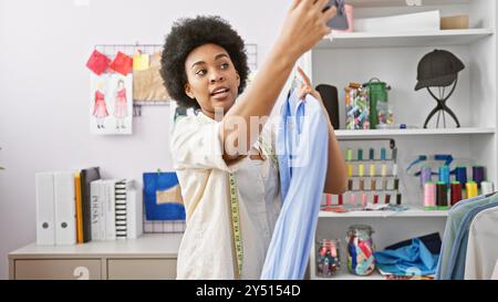 Femme afro-américaine tailleur prenant un selfie avec une chemise dans un atelier lumineux. Banque D'Images