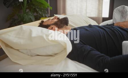 Homme barbu reposant paisiblement dans une chambre avec literie blanche et lumière naturelle douce. Banque D'Images