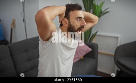 Homme barbu surpris dans le salon avec la main sur la tête, exprimant un choc ou un problème à l'intérieur. Banque D'Images