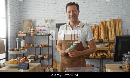 Bel homme hispanique dans un tablier debout dans une boulangerie avec du pain et des pâtisseries affichées en arrière-plan Banque D'Images