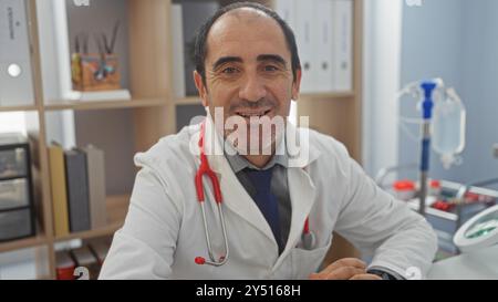 Médecin chauve hispanique d'âge moyen portant un manteau blanc et un stéthoscope rouge, souriant à la caméra dans une salle de clinique d'hôpital avec du matériel médical Banque D'Images
