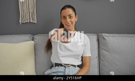 Femme regardant la télévision sur un canapé gris à l'intérieur tout en pointant la télécommande vers la caméra, portant une tenue décontractée Banque D'Images