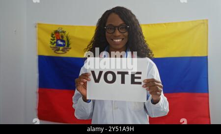 Une belle jeune femme afro-américaine aux cheveux bouclés se tient à l'intérieur devant un drapeau vénézuélien tenant un signe de vote, portant des lunettes et souriant Banque D'Images