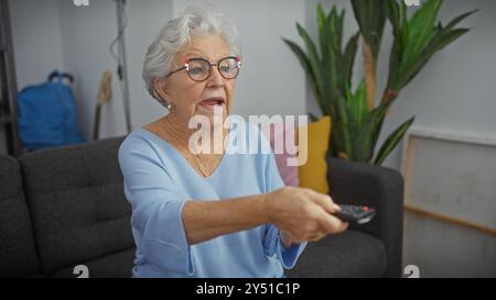 Femme âgée regardant la télévision confortablement dans son salon moderne tenant une télécommande Banque D'Images