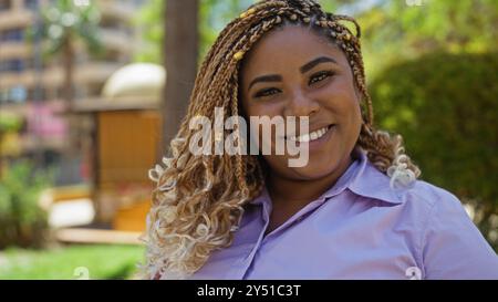 Une belle jeune femme afro-américaine avec des tresses sourit chaleureusement dans un cadre de parc urbain, mettant en valeur des paysages extérieurs vibrants dans la ville. Banque D'Images