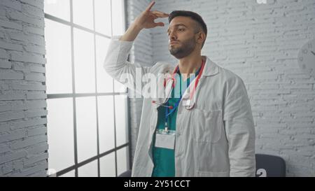 Jeune médecin hispanique barbu dans une salle d'attente d'hôpital, portant un manteau blanc et un stéthoscope, regarde soigneusement par la fenêtre. Banque D'Images