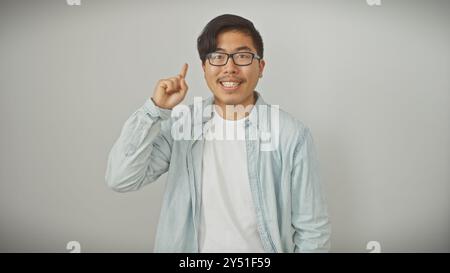 Souriant jeune homme asiatique dans des lunettes faisant un point avec le doigt levé, habillé décontracté sur fond blanc isolé. Banque D'Images