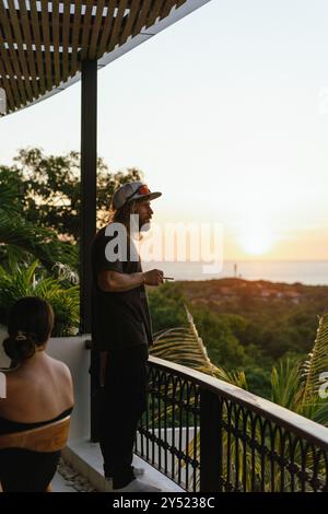 Un homme en vacances, fumant sur la terrasse au coucher du soleil. Banque D'Images